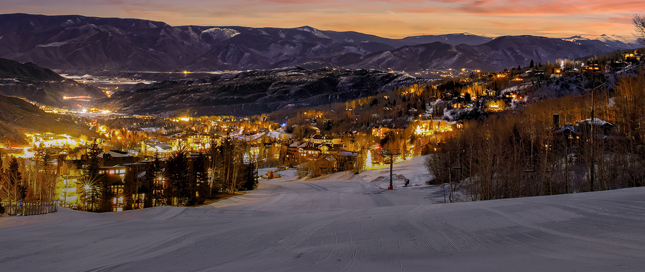 Snow-covered ski slope overlooking Aspen, Colorado, with illuminated mountain homes and a scenic mountain range at sunset.